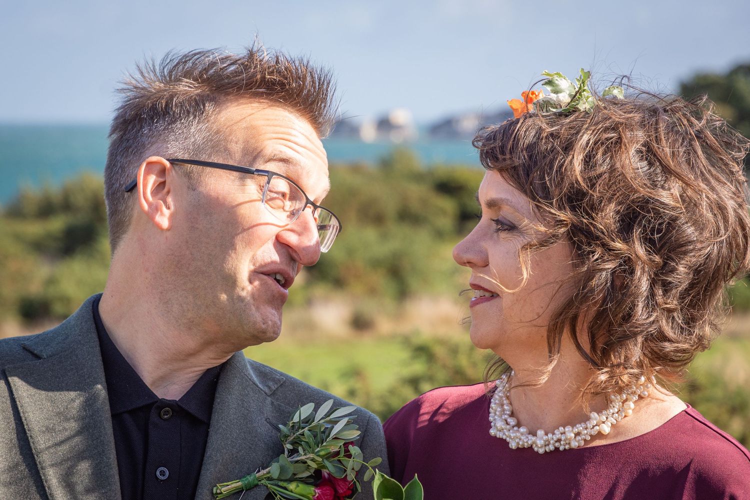 Couple gazing at each other outdoors, man in glasses holding red roses, woman with floral hair accessory, ocean in background.