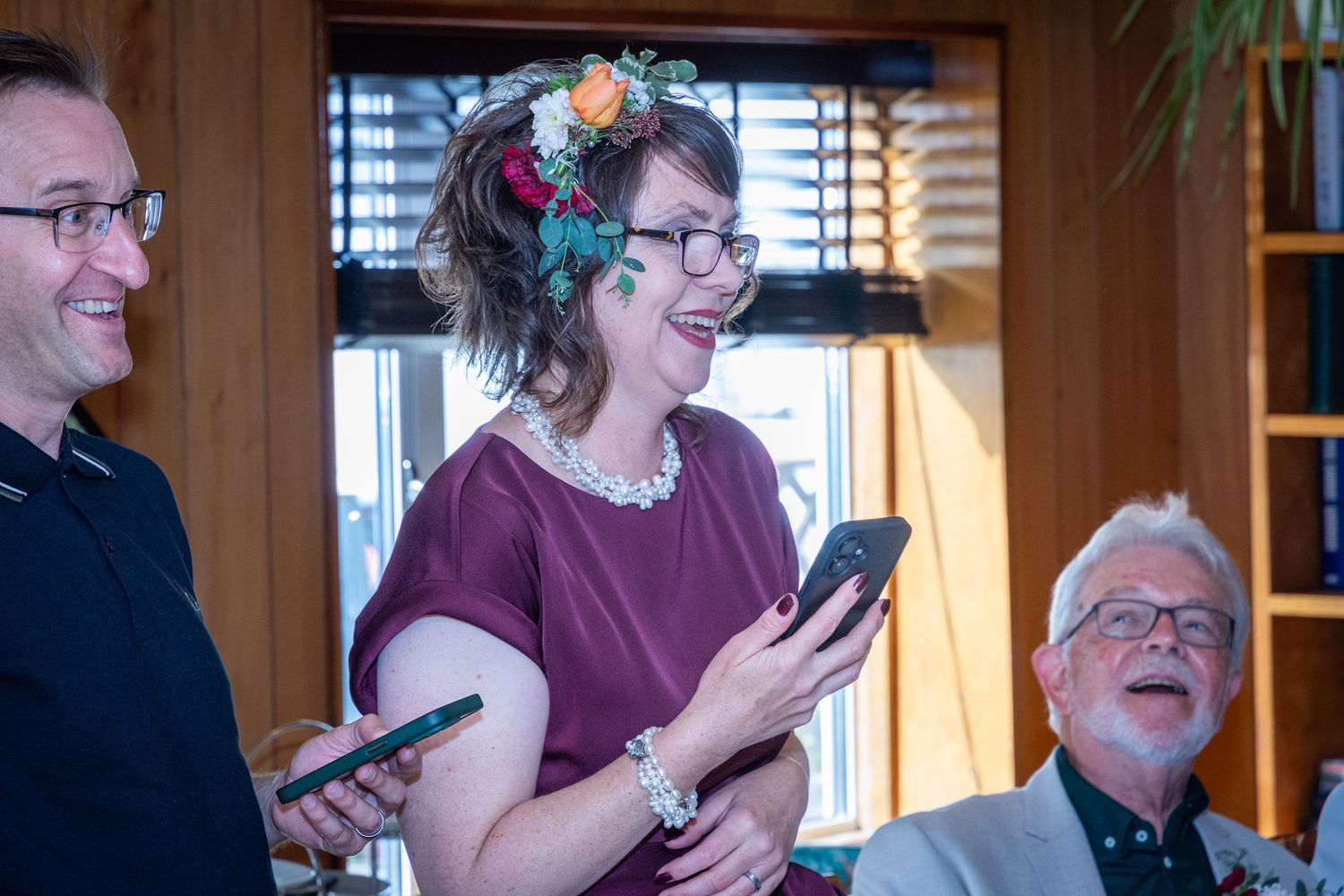 Woman with flower crown laughs holding phone at indoor wedding celebration with guests.