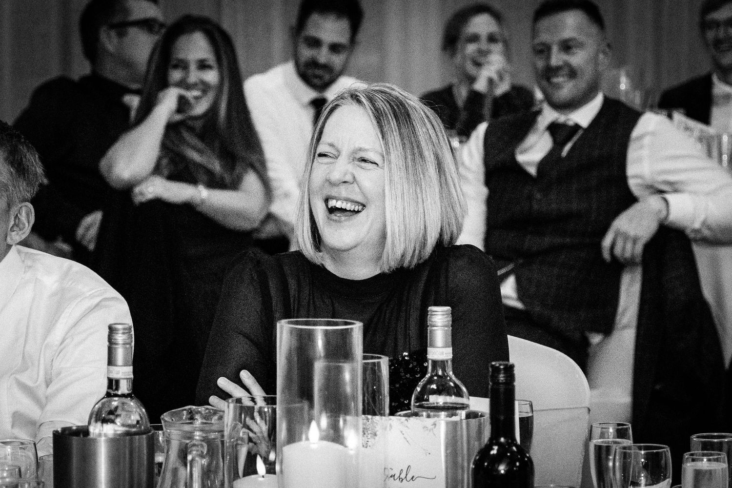Black and white photo of a laughing woman at a wedding reception dinner table with guests celebrating in background.