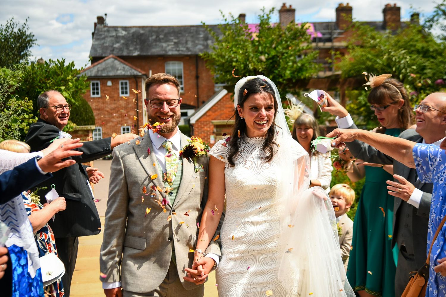 Bride and groom walk hand in hand through confetti shower surrounded by guests outside brick venue.