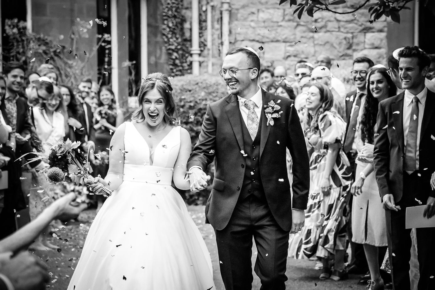 Bride and groom walk through confetti shower surrounded by cheering guests at outdoor wedding ceremony.