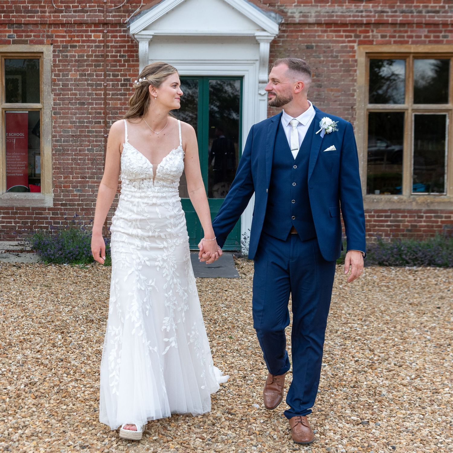 A bride in a white lace gown and groom in a navy suit hold hands walking outside a brick venue.