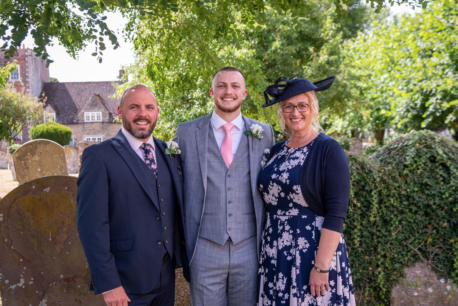Three wedding guests pose outdoors near gravestones, man in grey suit with pink tie flanked by two smiling attendees.