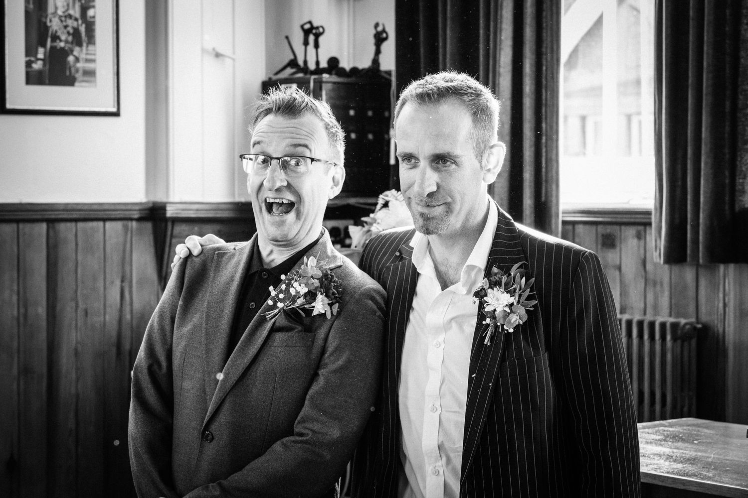 Two men in suits with boutonnieres smiling together at a wedding celebration in black and white photo.