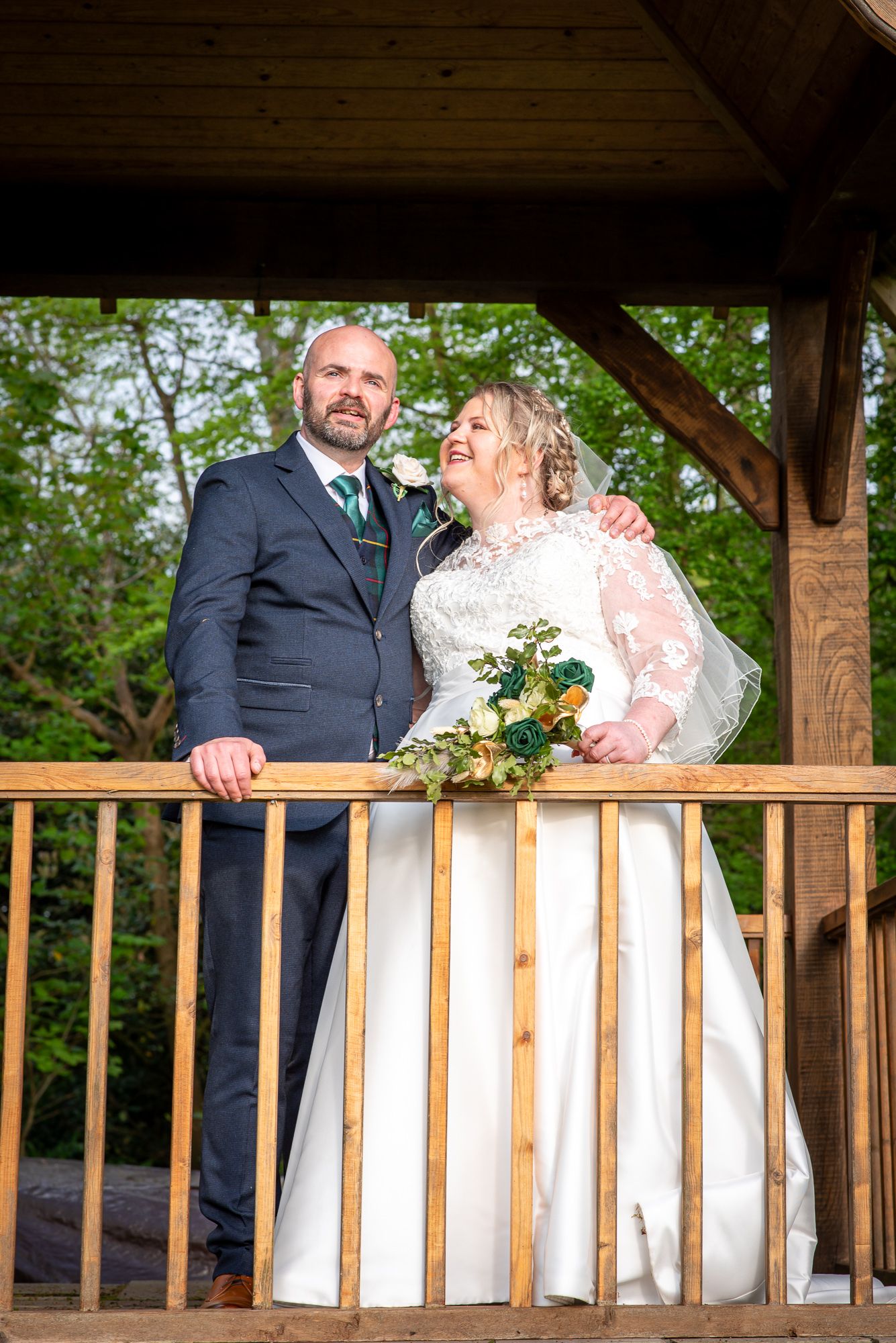 Bride and groom standing on wooden gazebo balcony, bride in lace dress holding bouquet, groom in navy suit.