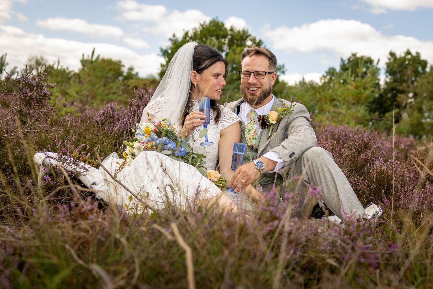 Bride and groom sitting in purple heather fields, smiling together on their wedding day outdoors.