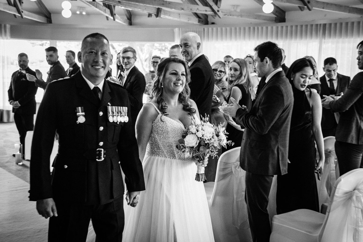 Black and white photo of a smiling military groom and bride walking down the aisle at their wedding ceremony.