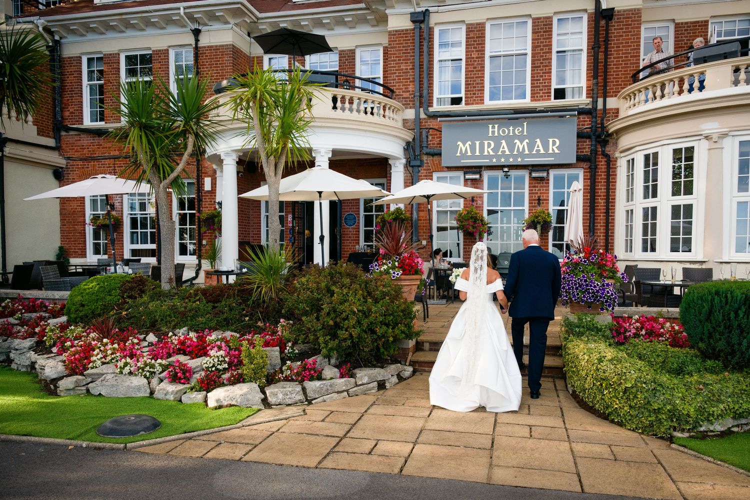 Bride and groom walking toward Hotel Miramar entrance surrounded by colorful gardens and palm trees on their wedding day.