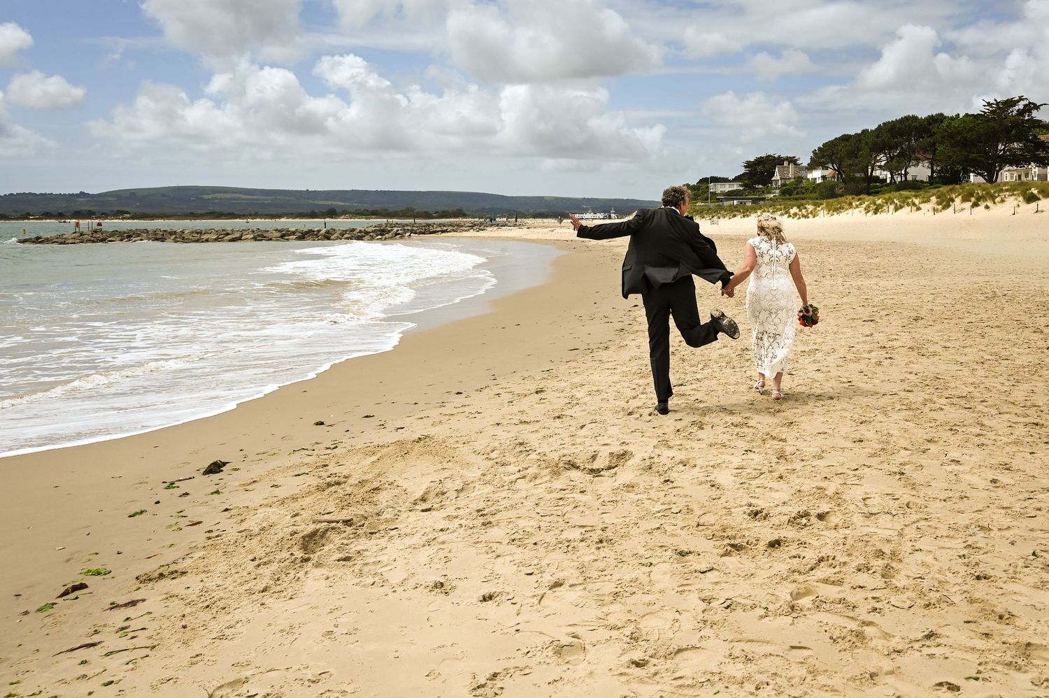 Bride and groom walking hand in hand along a sandy beach on their wedding day.