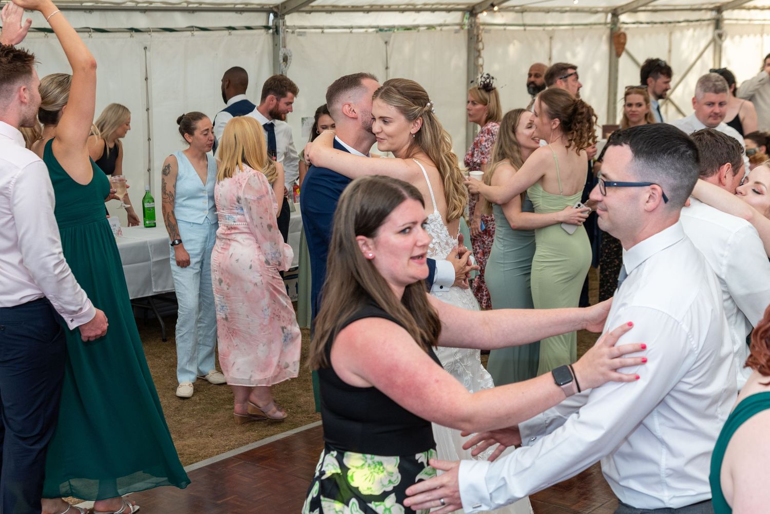 Guests dance and celebrate on the dance floor at a lively wedding reception under a marquee tent.