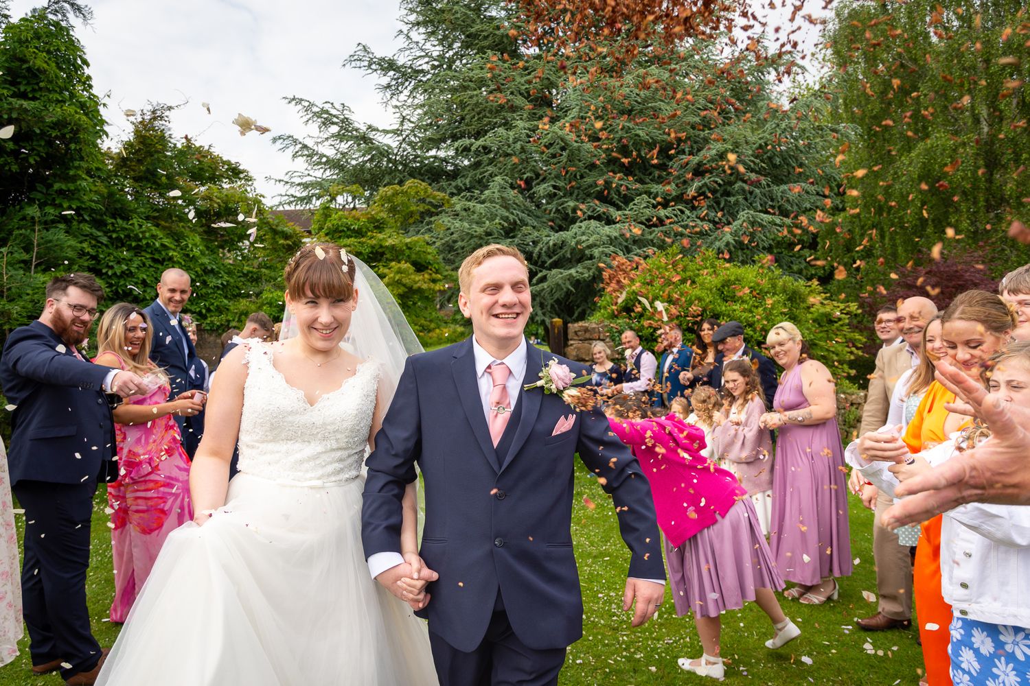 Wedding party and guests gathered outdoors for a celebratory photo on a garden path.