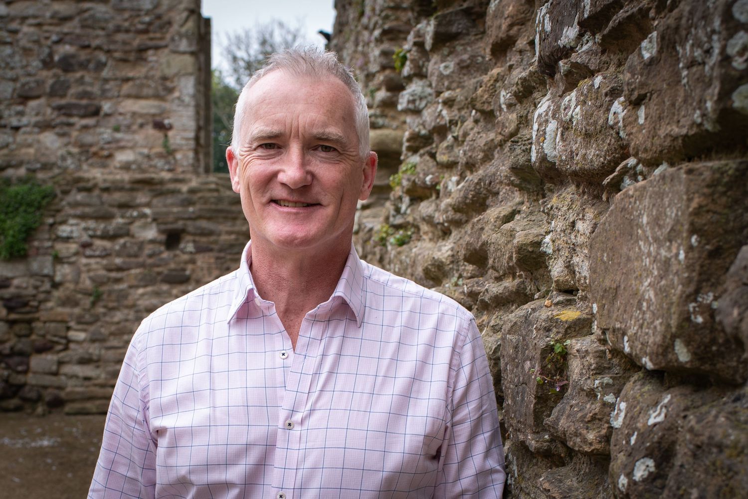 Smiling older man in pink checked shirt leaning against rustic stone wall at historic ruins.