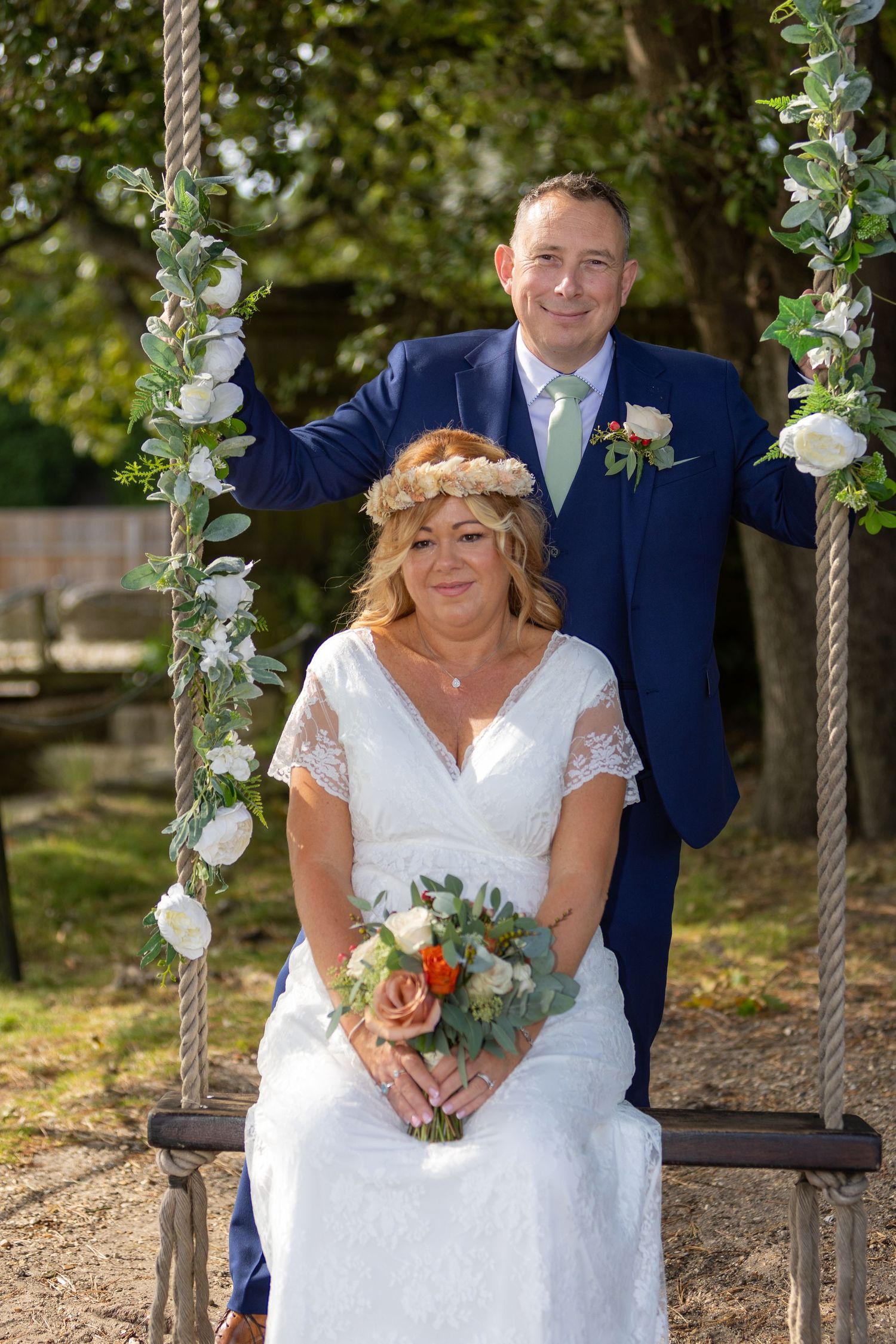 Bride in white lace dress with flower crown sits on swing holding bouquet, groom in navy suit stands behind her outdoors.