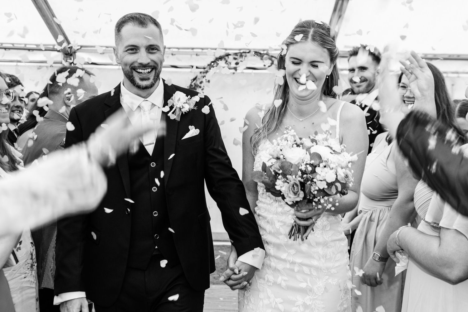 Happy wedding couple walks down aisle as guests throw white flower petals during joyful recessional celebration.
