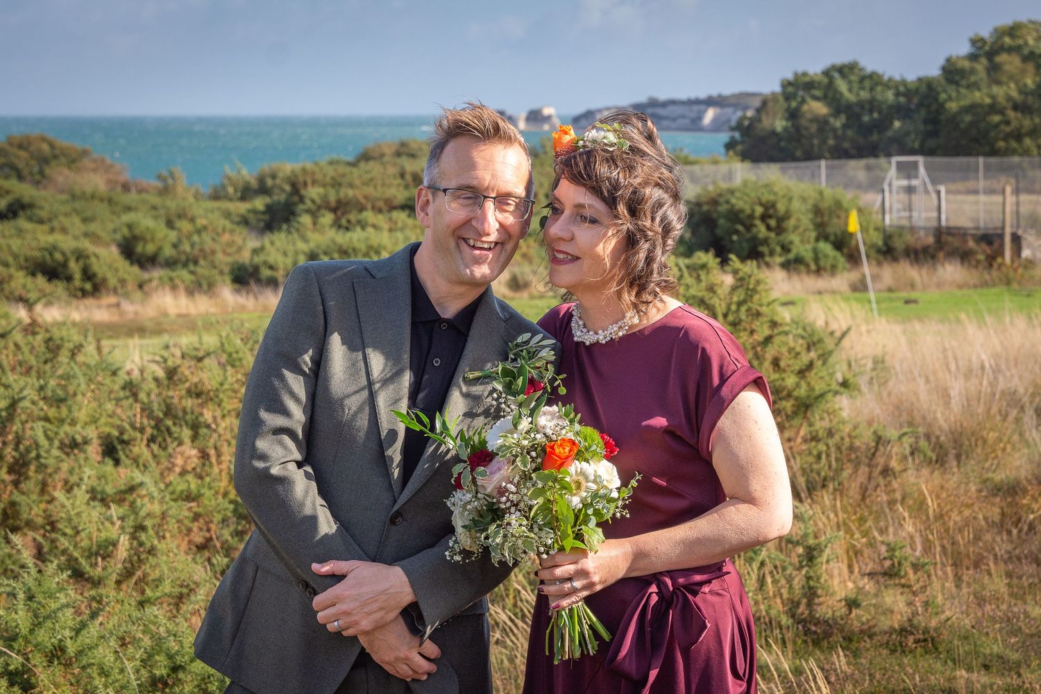 A couple in formal attire poses together outdoors against a coastal landscape during a special occasion.