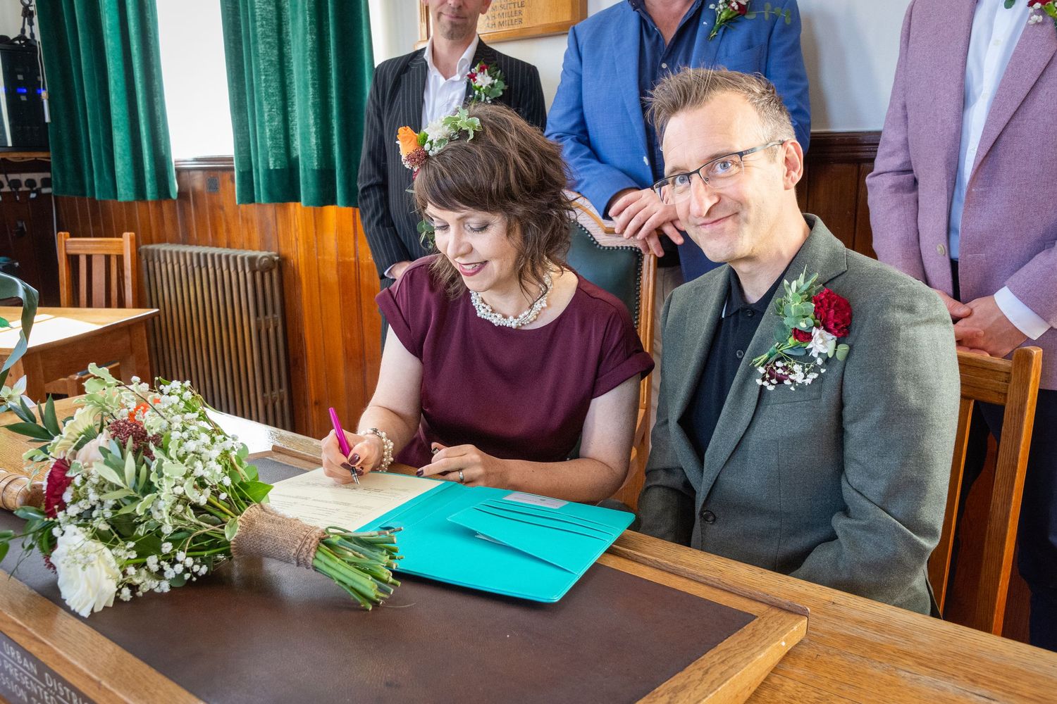 Bride and groom signing wedding register at a wooden desk with a teal folder during their ceremony.