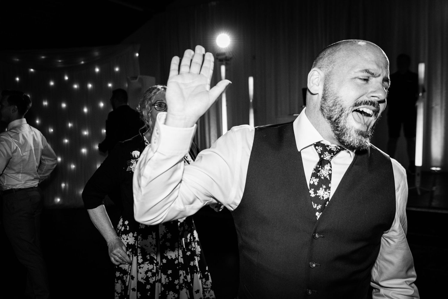 Bald bearded man dancing joyfully at a wedding reception, raising his hand and laughing in black and white photo.