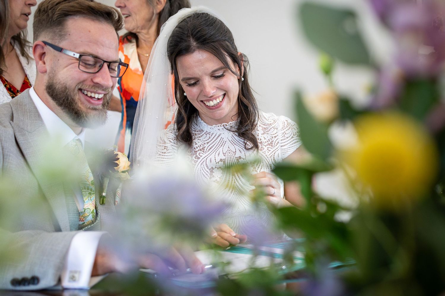 Happy couple smiling during wedding ceremony signing, surrounded by colorful flowers in soft focus foreground.