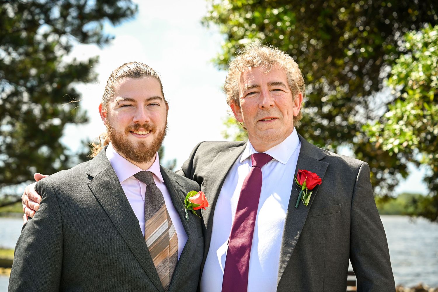 Two men in suits with boutonnieres posing outdoors near water at a formal event.