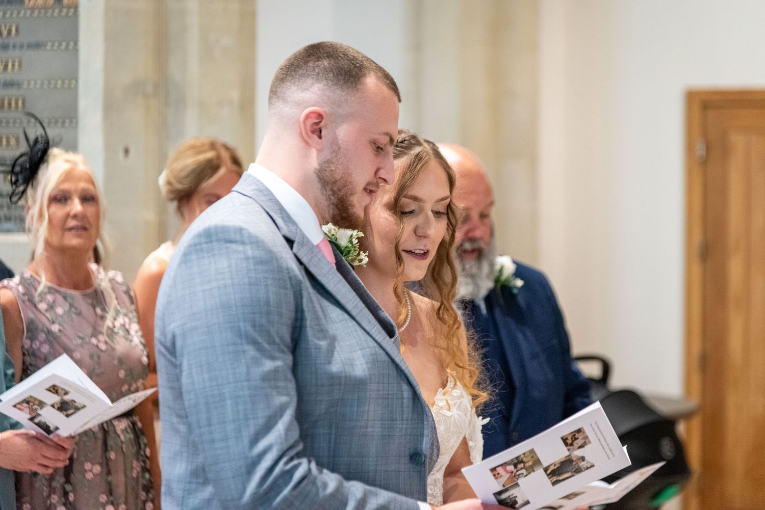 Bride and groom share emotional moment reading wedding program during church ceremony with guests seated behind.
