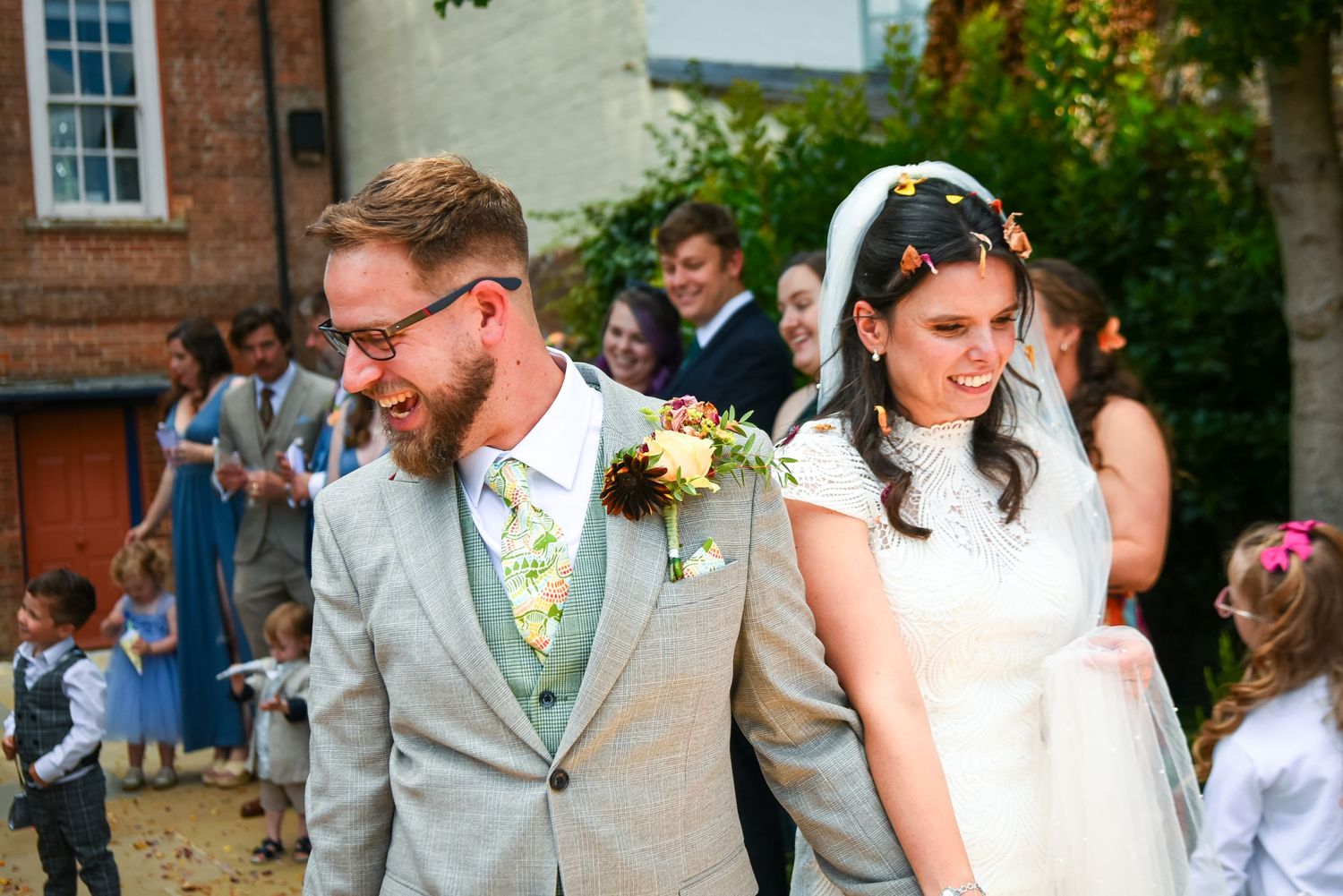 Smiling bride and groom walking through confetti shower at outdoor wedding celebration with guests cheering.