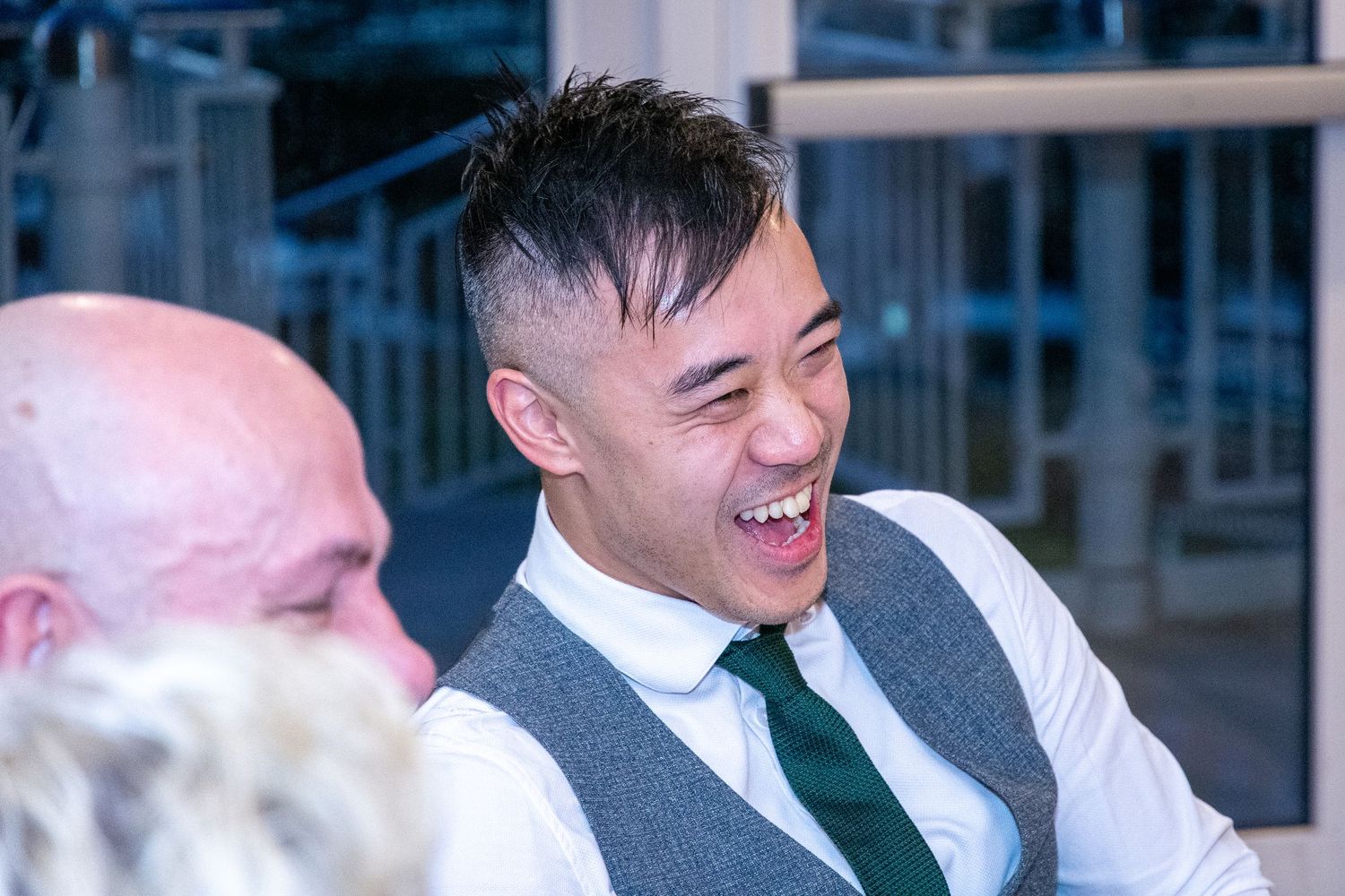Man in grey vest and green tie laughing joyfully at a social gathering with others nearby.