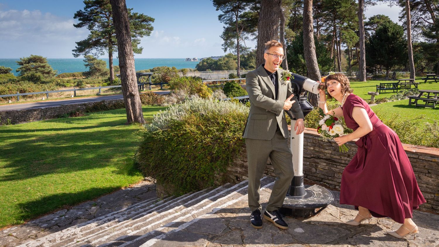 Couple in formal attire posing in a scenic garden with ocean views and tall pine trees on a sunny day.