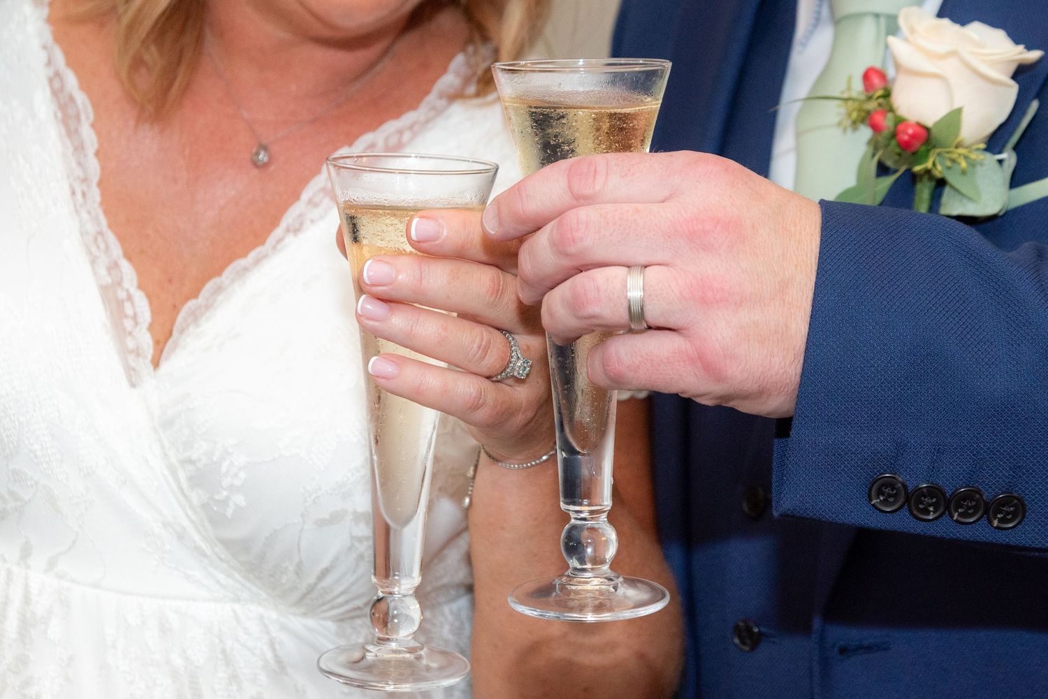 Newlywed couple toasting with champagne flutes, wearing wedding rings, bride in white dress, groom in navy suit.