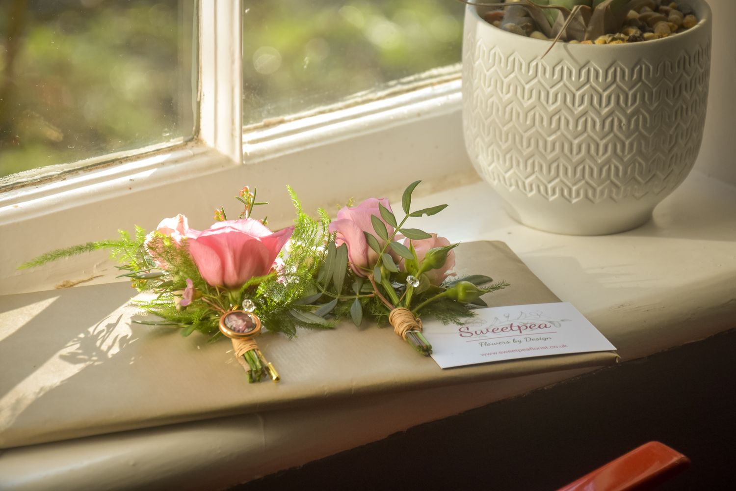 Pink rose boutonnieres with greenery on a book beside a succulent plant on a sunny windowsill.