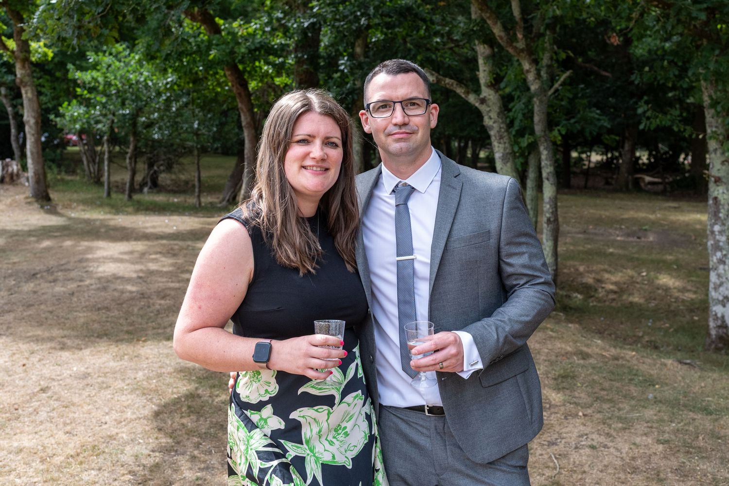 A smiling couple holding drinks outdoors at a garden event, dressed in smart casual attire among lush green trees.