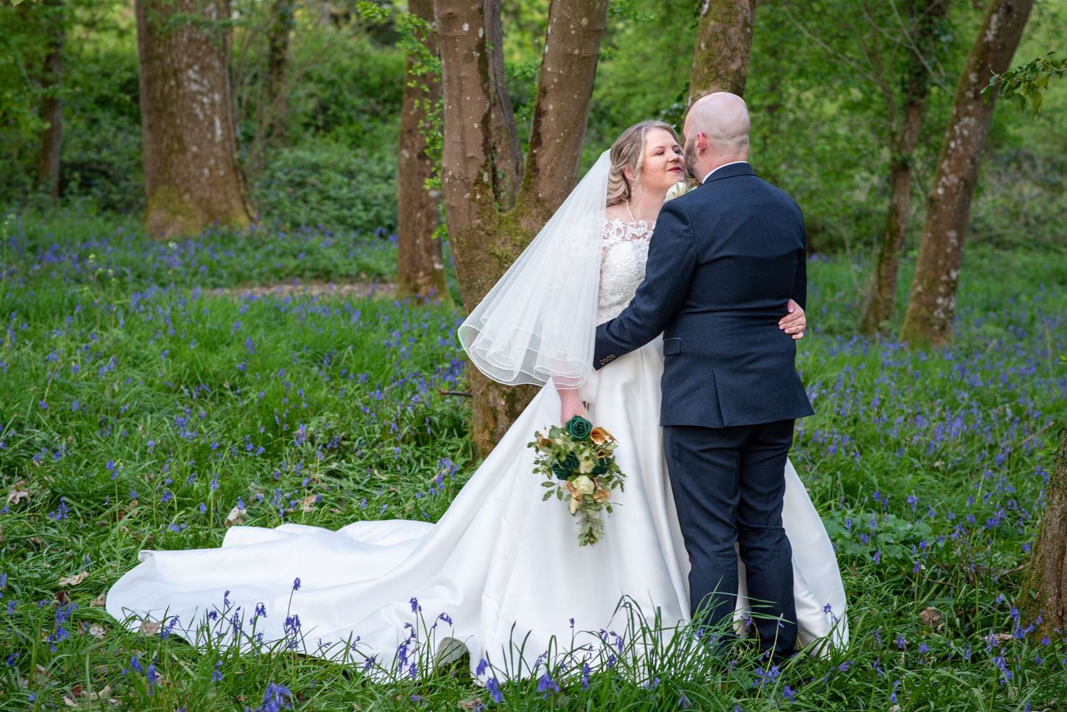 Bride and groom embrace in a bluebell woodland, bride wearing a white gown with veil and holding a bouquet.