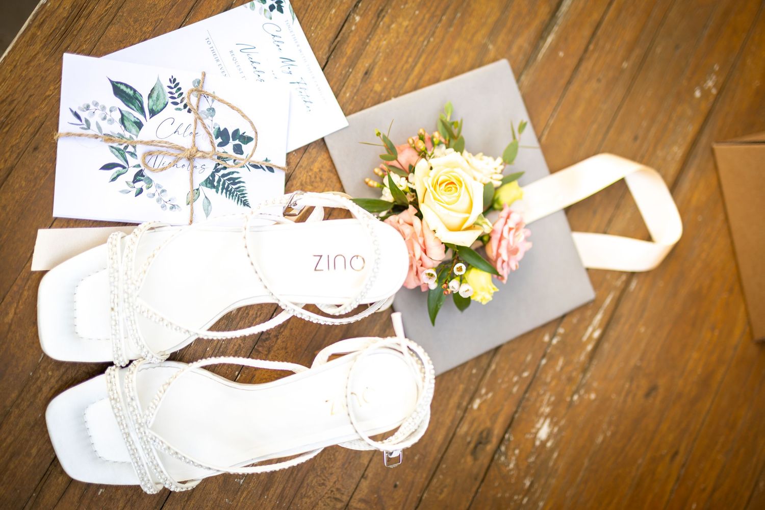 White ZINO bridal sandals displayed with wedding invitation and pink floral bouquet on rustic wood floor.