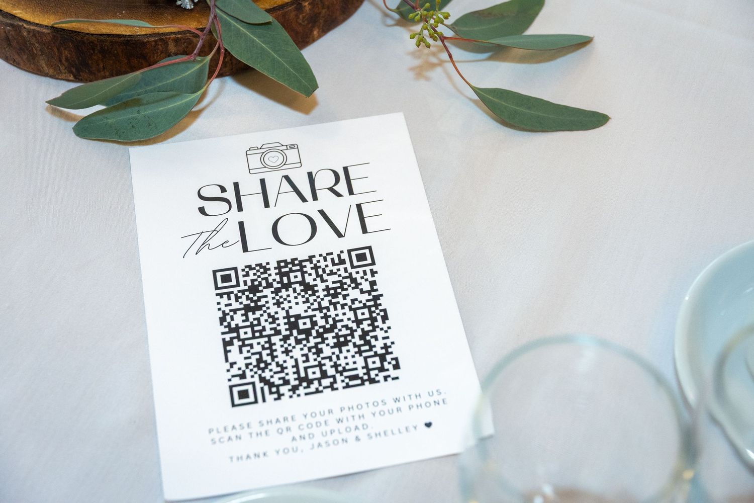 Wedding Share The Love QR code card displayed on a decorated reception table with greenery.