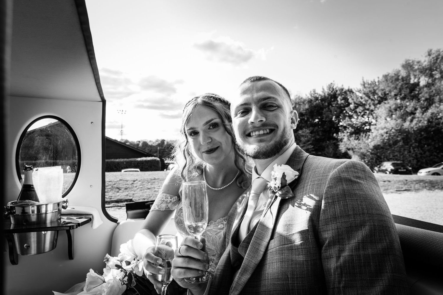 Black and white wedding photo of smiling bride and groom holding champagne glasses inside a boat.