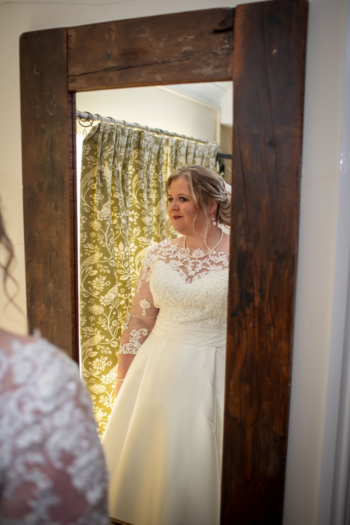 Bride in lace wedding dress admiring her reflection in a rustic wooden mirror with floral curtains.