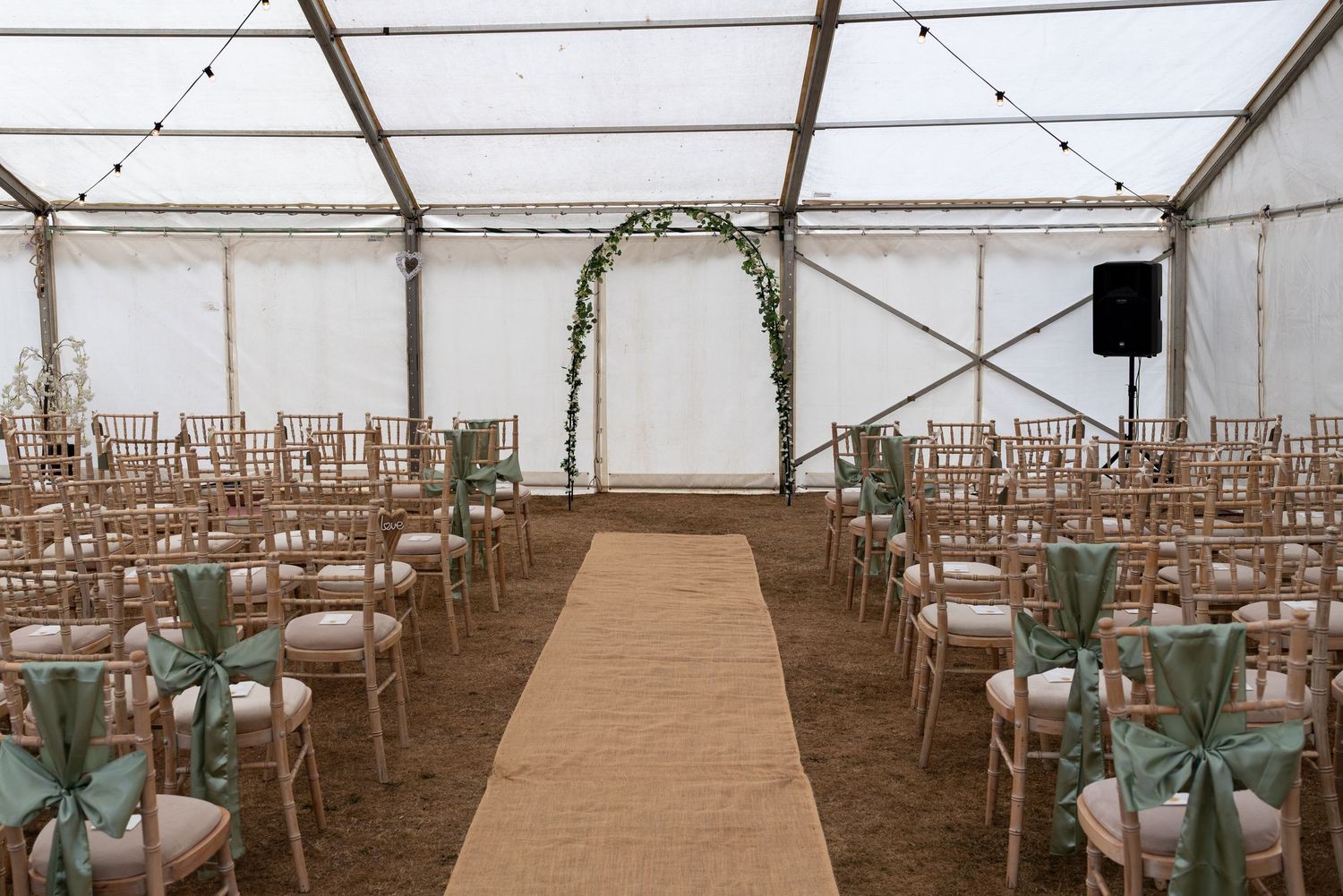 Elegant wedding ceremony tent with chiavari chairs, sage green ribbon bows, burlap aisle runner, and floral arch at the altar.