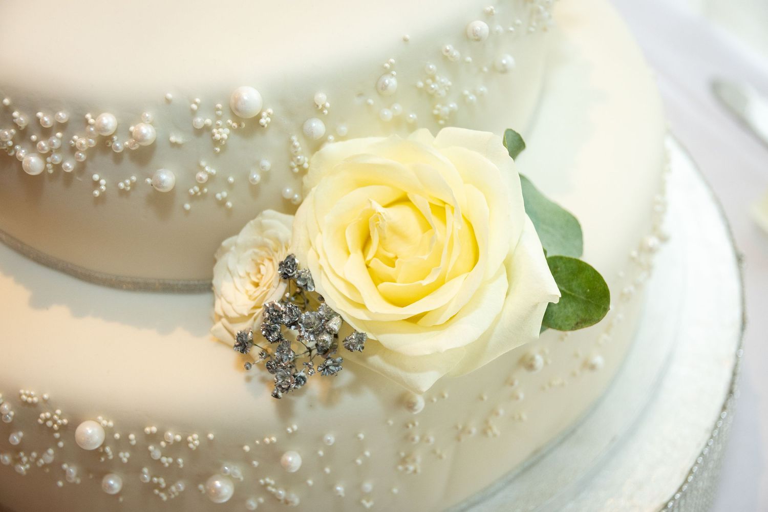 White wedding cake with pearl details and a yellow rose, small flowers, and green leaves decoration.