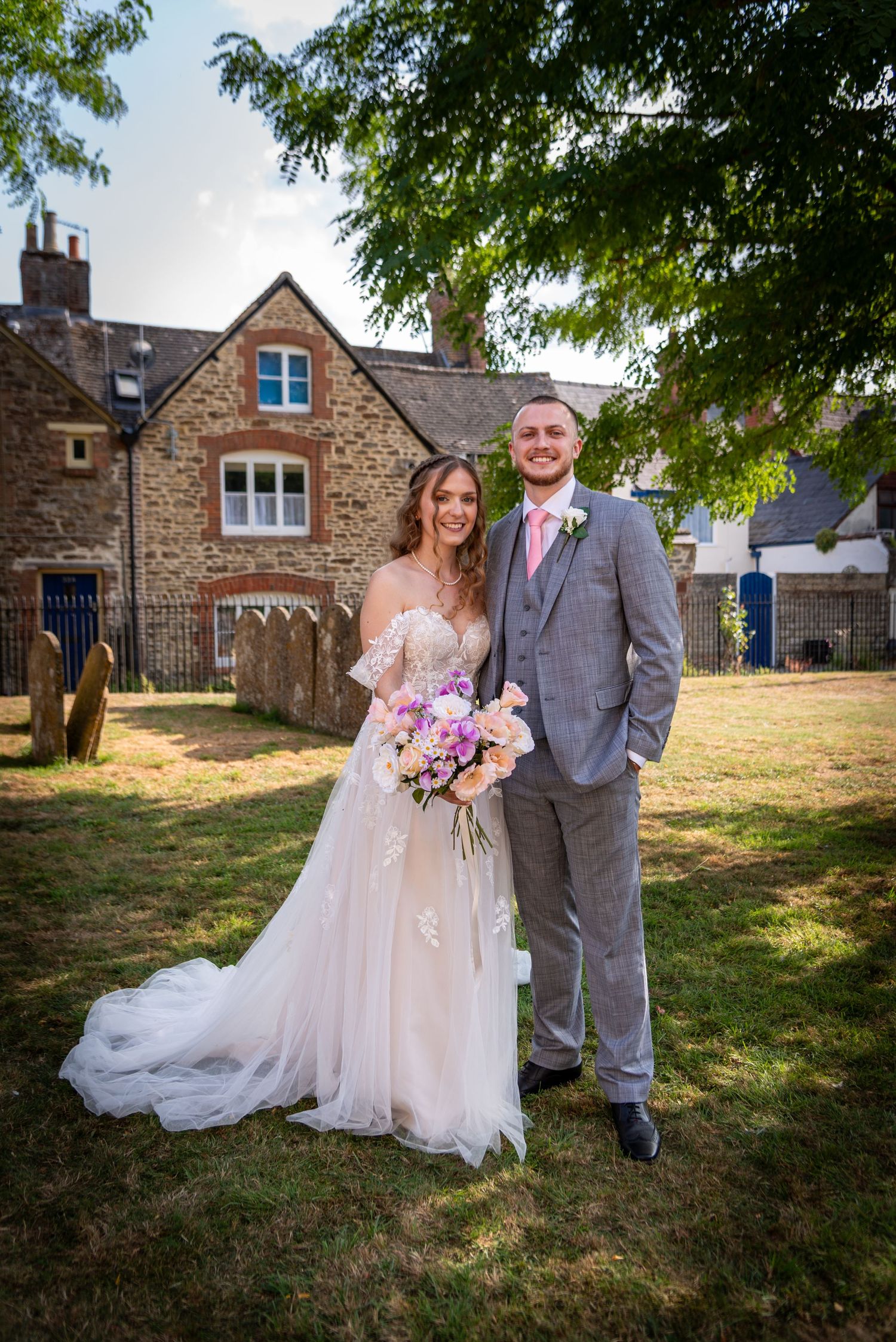 Bride and groom posing together outdoors on their wedding day, with a stone building backdrop and lush greenery.