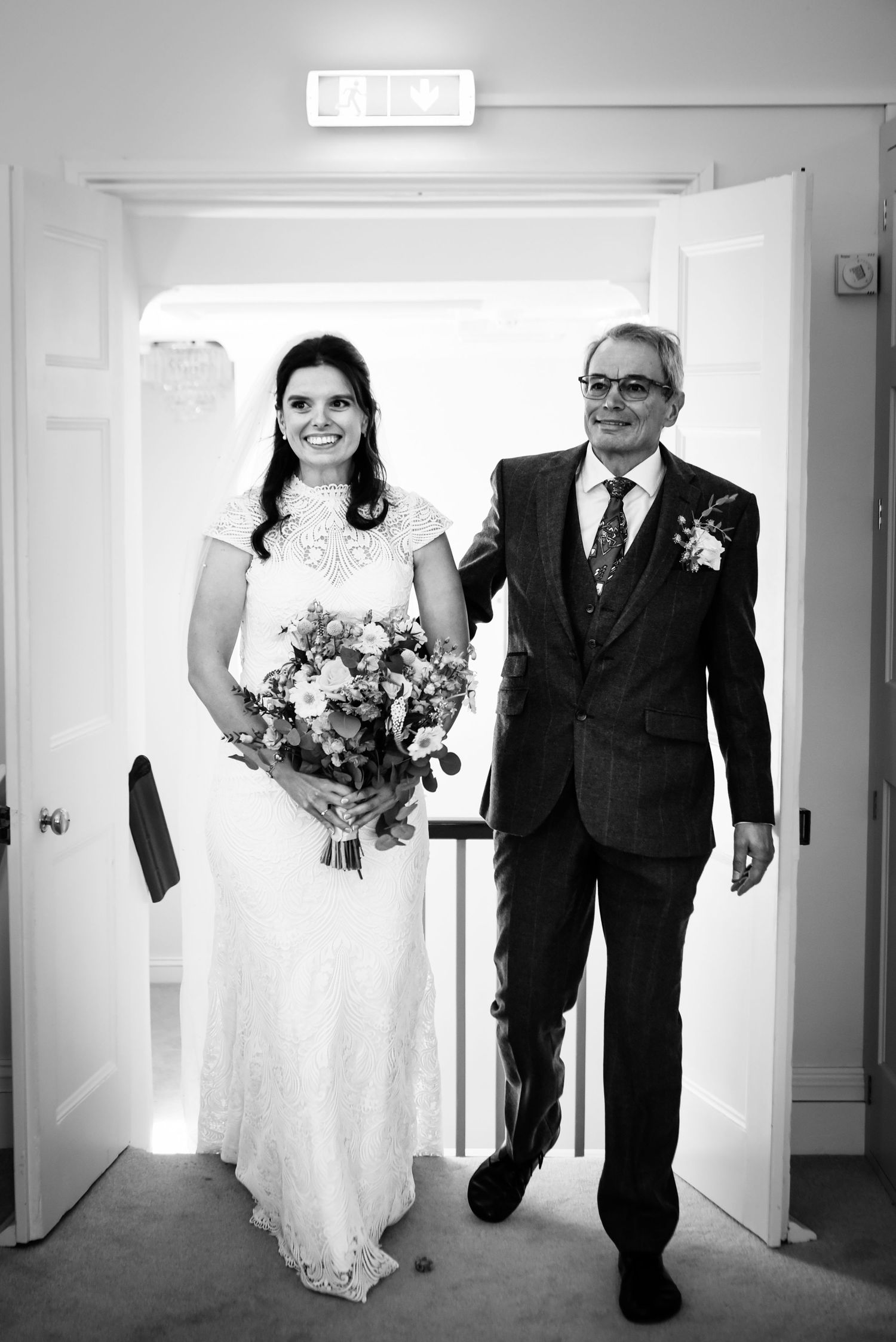 Bride in white lace gown holding bouquet walks with suited older man through doorway in black and white wedding photo.