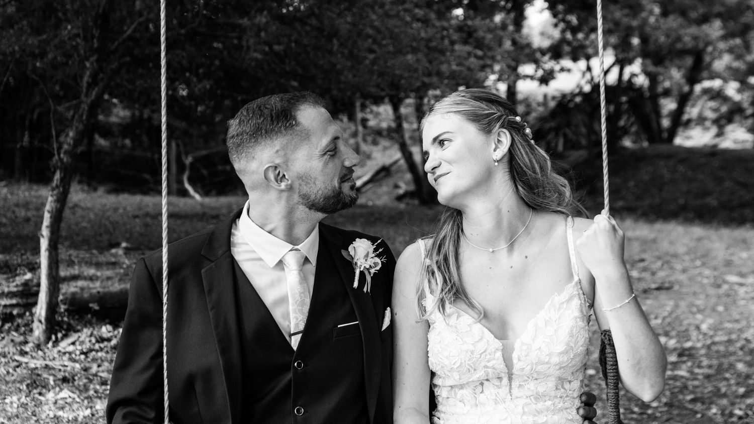 Black and white wedding photo of bride and groom sharing a romantic moment on a swing surrounded by trees.
