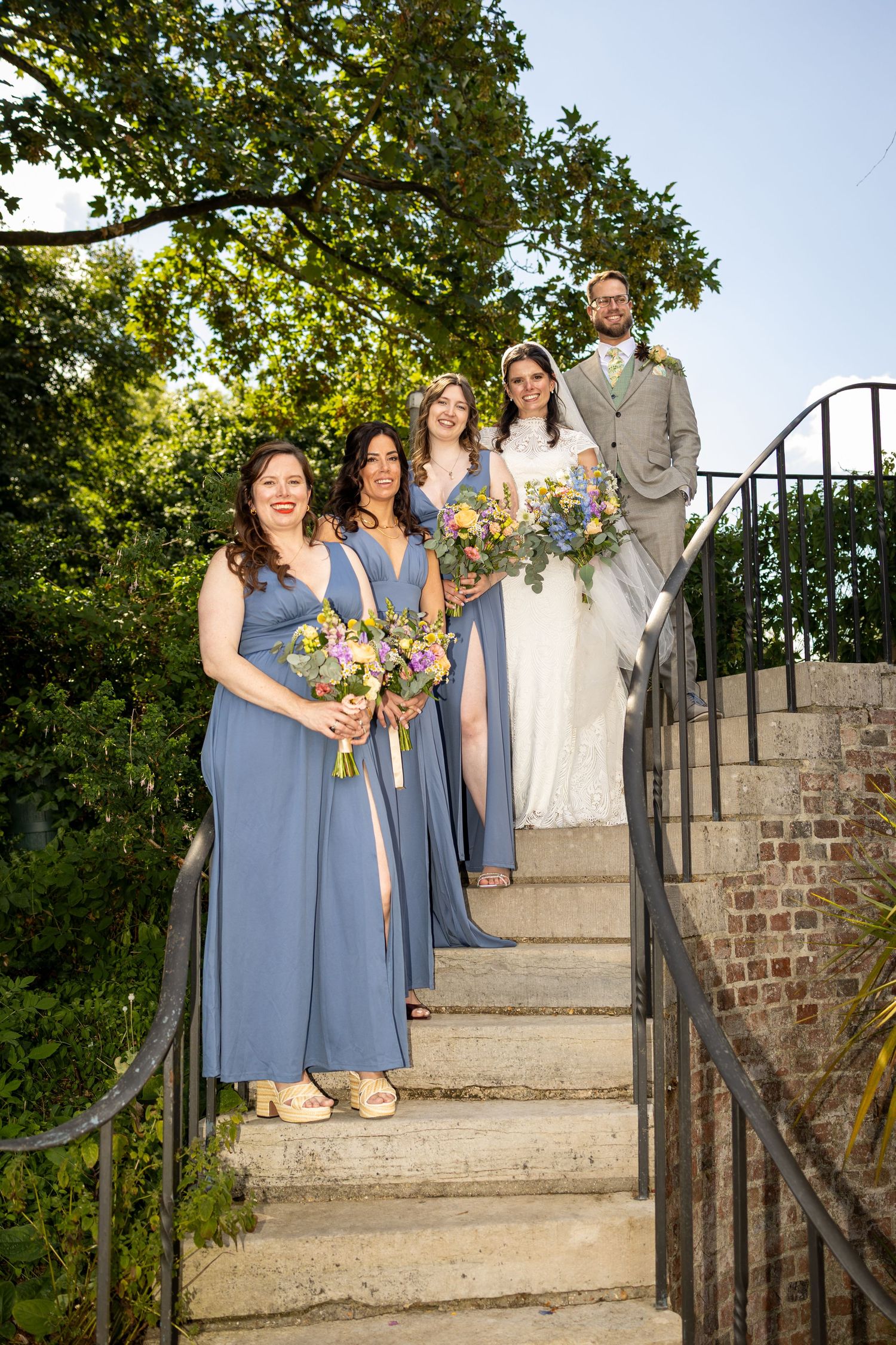 Wedding party poses on brick staircase with bridesmaids in blue gowns holding colorful bouquets beside bride and groom.
