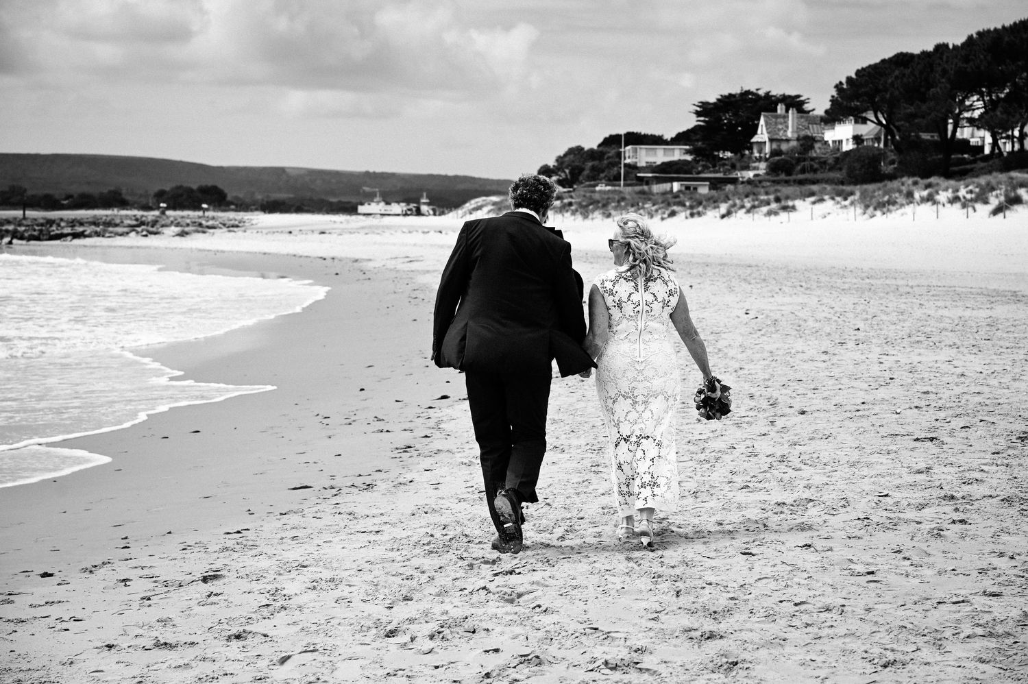 Black and white photo of a bride and groom walking hand in hand along a sandy beach on their wedding day.