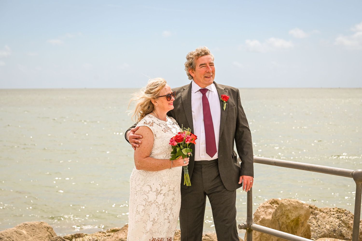 Bride and groom posing by the seaside on their wedding day, with the bride holding a red bouquet.