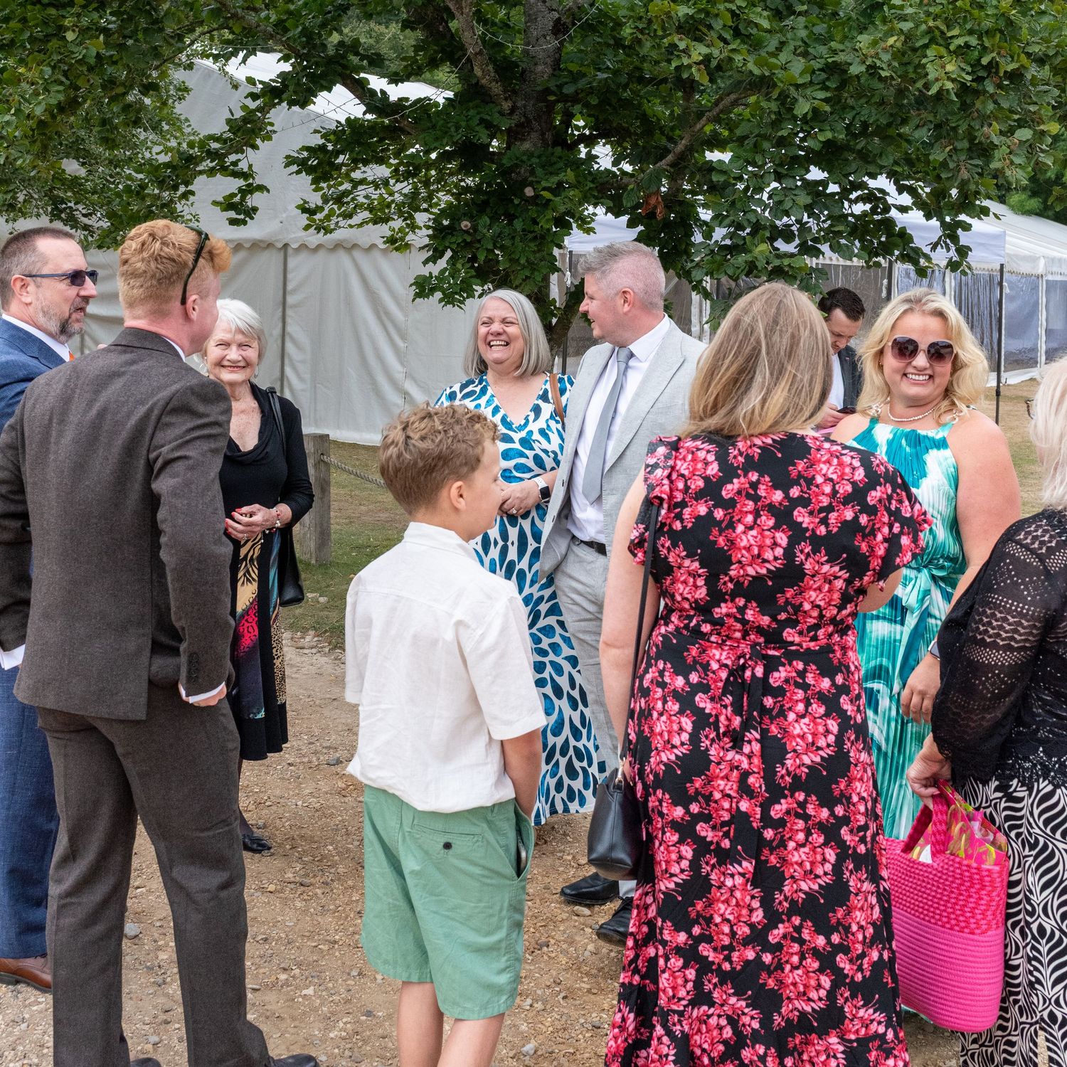 Guests in formal attire mingle outdoors at a summer wedding reception near a marquee tent surrounded by trees.