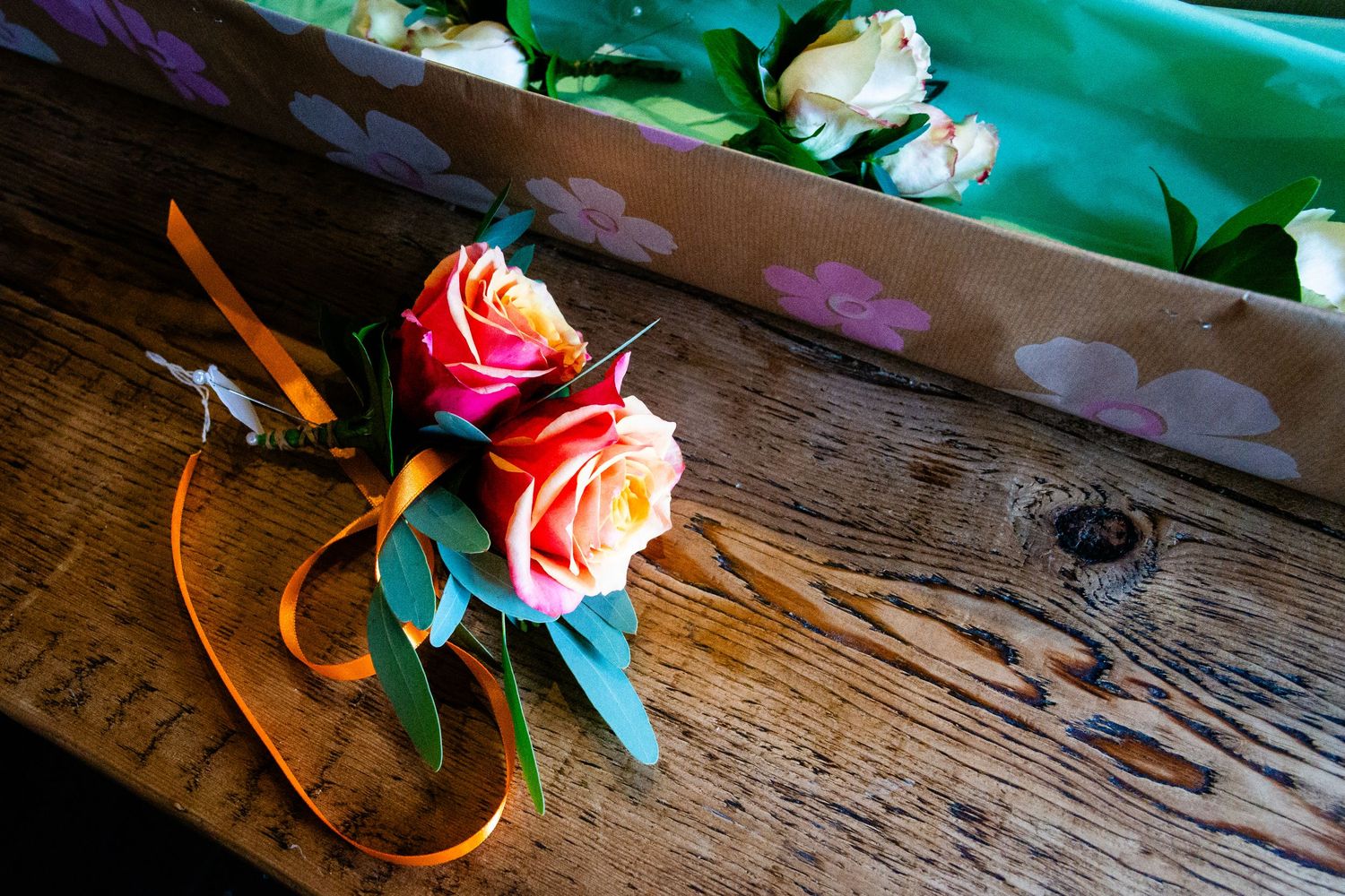 Two peach roses with orange ribbon and greenery on rustic wooden table, with wrapped flowers in background.