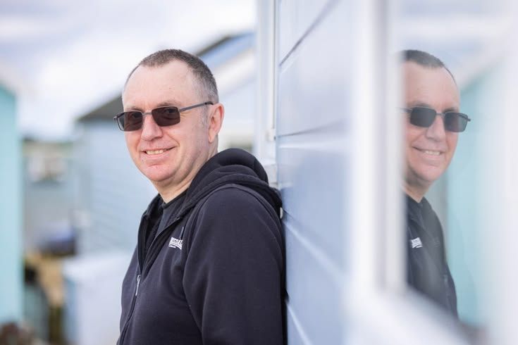 Middle-aged man in black hoodie and sunglasses leaning against a wall, with his reflection visible in a window.