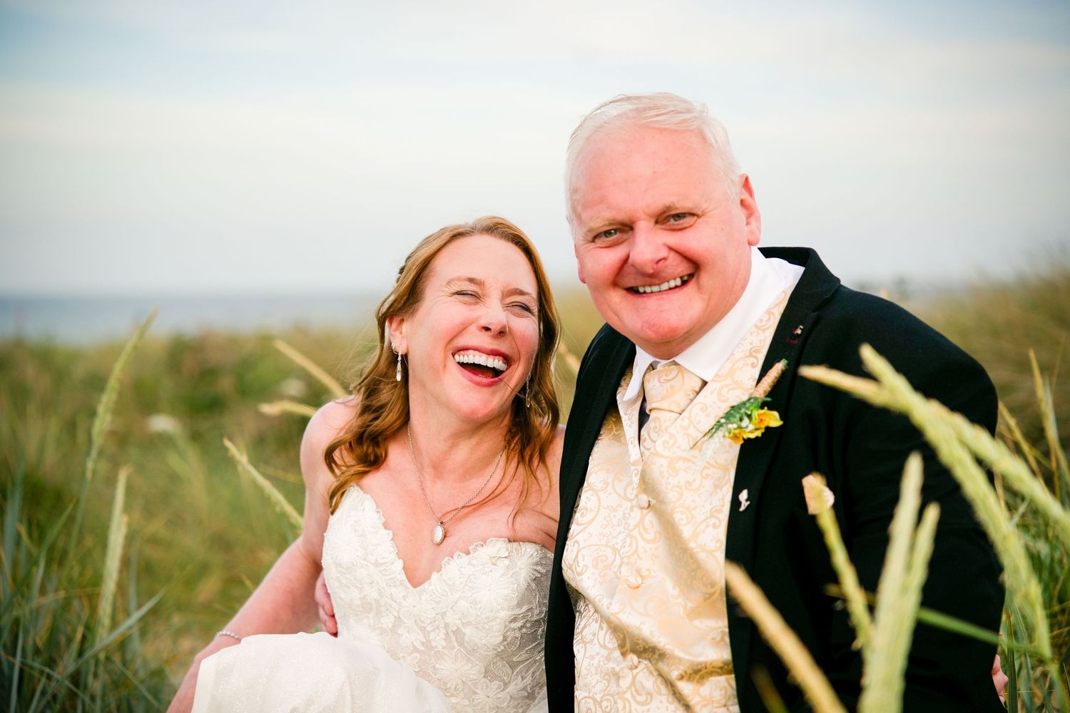 Happy bride and groom laughing together outdoors near beach grass on their wedding day.