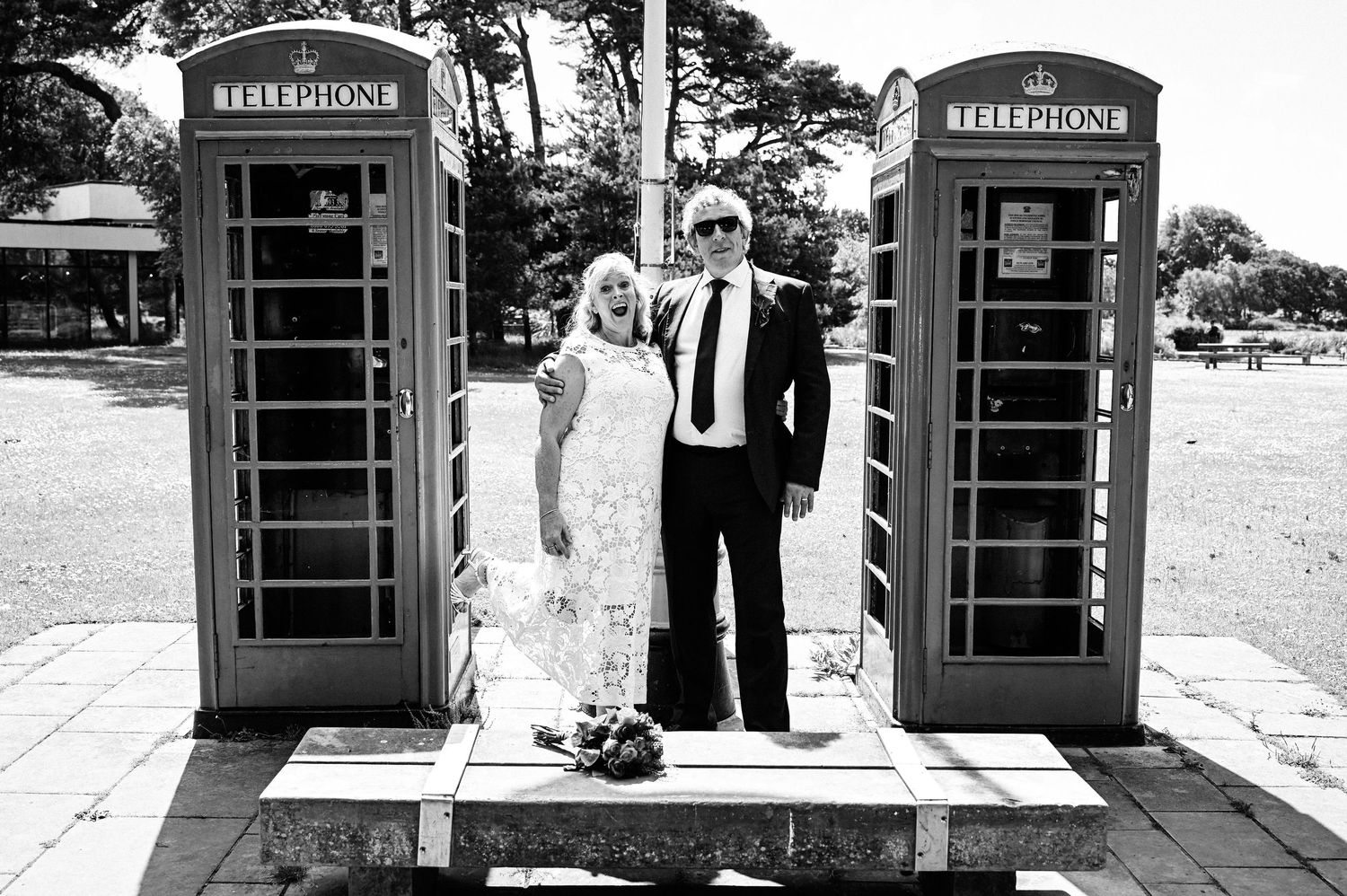 Couple in wedding attire posing between two classic British red telephone boxes in black and white photo.