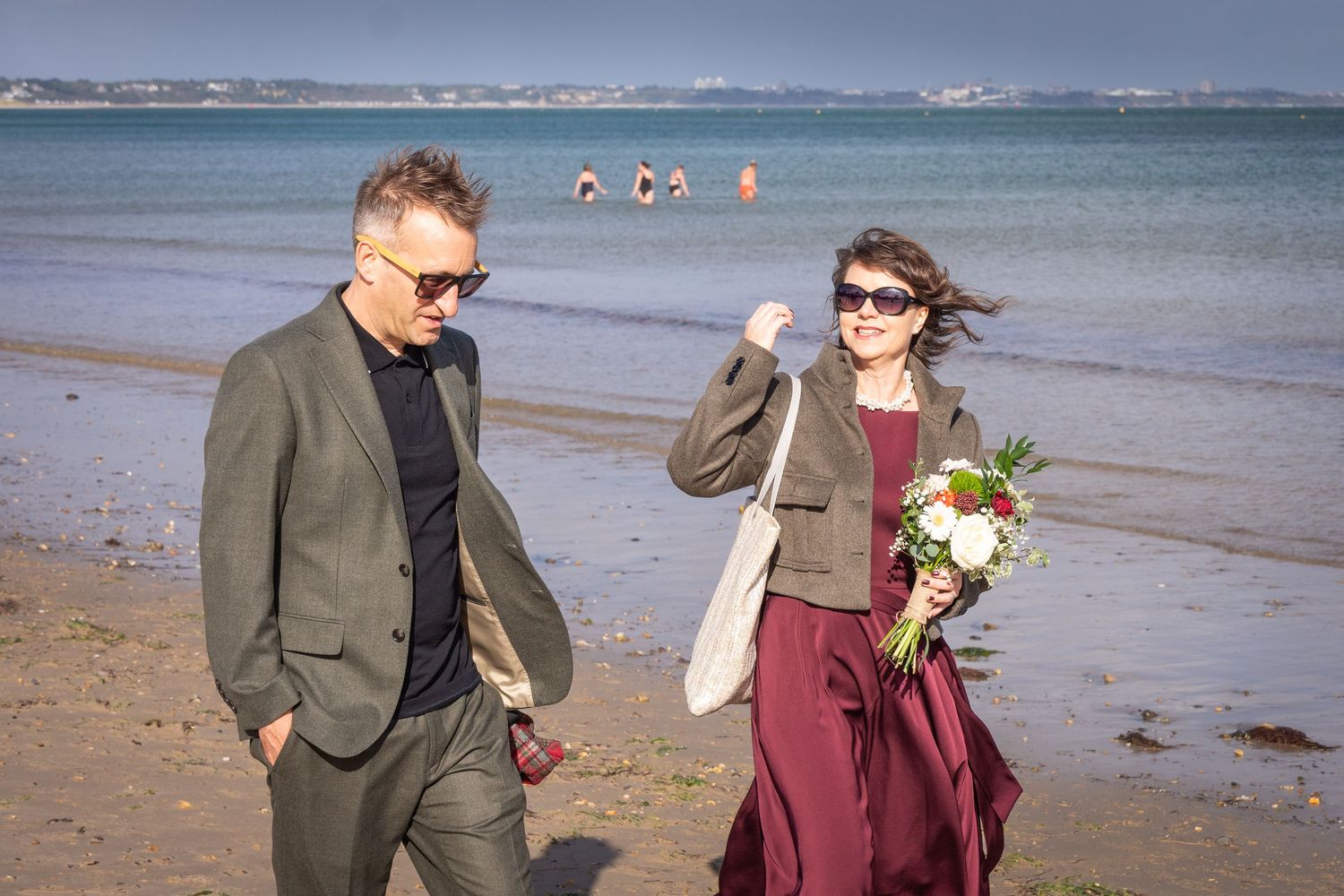 Couple walking on a sandy beach, woman holding a bouquet, man in suit, ocean in background on a sunny day.