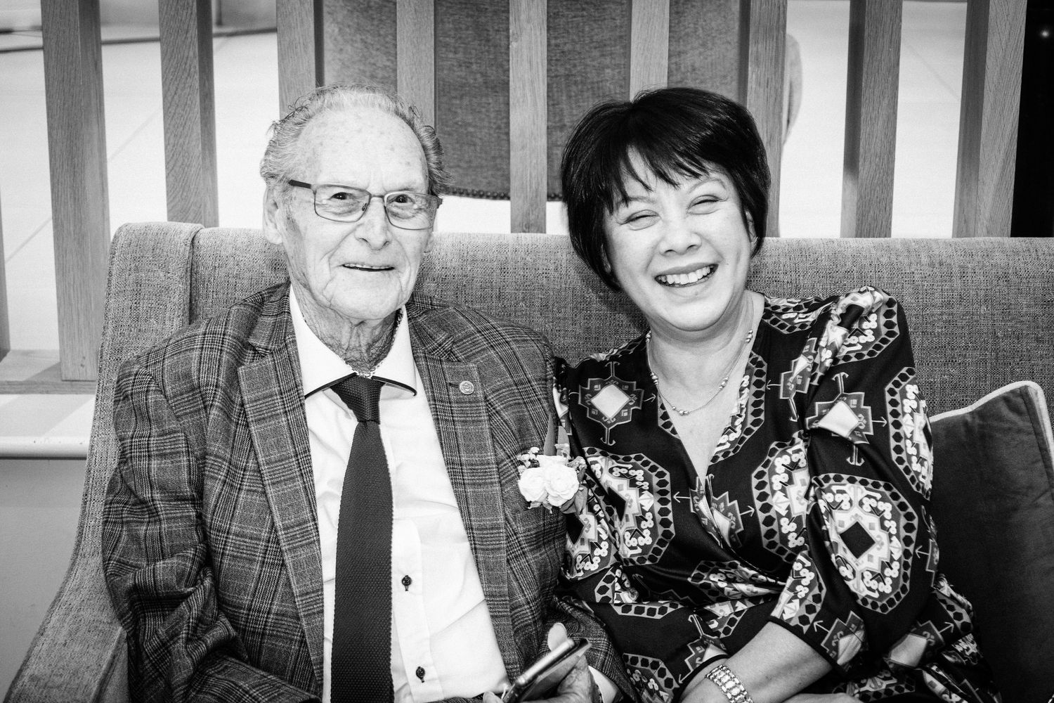 Elderly man in plaid suit and woman in patterned dress smiling together on a sofa in a black and white photo.
